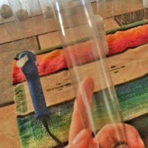 Puede incluir: Un tubo de vidrio transparente con una tapa blanca en un extremo. El tubo está sobre una mesa de madera con una manta colorida debajo. Hay varios tubos de vidrio más pequeños en el fondo.