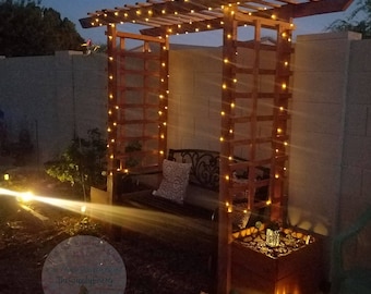 Könnte beinhalten: Eine Holzpergola mit Lichterketten, die darüber drapiert sind. Die Pergola befindet sich in einem Garten mit einer Bank und einem Blumenkasten. Die Lichter sind an und erzeugen ein warmes Licht.