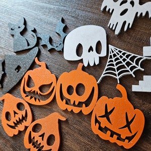 Op de afbeelding: Een verzameling Halloween-decoraties gemaakt van vilt. De decoraties omvatten een ​​spook, een schedel, een spinnenweb, een grafsteen en verschillende pompoenen met verschillende gezichtsuitdrukkingen.