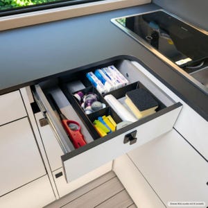 Könnte beinhalten: Eine offene Küchenschublade mit schwarzen Trennelementen, die ein rotes Feuerzeug, mehrere kleine metallische Kaffeekapseln, weiße rechteckige Schachteln, eine weiße Kunststoffrolle, gelbe und blaue Kunststoffklammern sowie einen gelb-schwarzen Schwamm enthält.