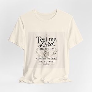 Può includere: Una t-shirt a maniche corte color crema con una stampa grafica nera sul petto. Il design include il testo "Test me, Lord, and try me, examine my heart and my mind" con un'illustrazione centrale di un cuore anatomico e "Psalms 26:2".