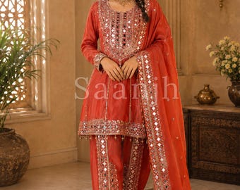 Peut inclure: Un ensemble orange vif composé d'une tunique à manches longues avec une broderie complexe en miroir sur l'encolure et l'ourlet, assortie d'un pantalon style dhoti et d'une dupatta transparente ornée de miroirs drapée sur une épaule.