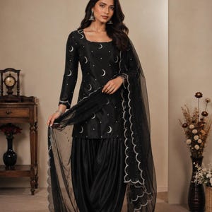 Op de afbeelding: Een zwarte salwar kameez-set bestaande uit een tuniek met lange mouwen, een vierkante halslijn en een patroon van zilveren maansikkels en stippen. De outfit bevat een bijpassende zwarte geplooide broek en een transparante zwarte sjaal.