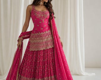 Könnte beinhalten: Eine leuchtend fuchsiafarbene ärmellose Tunika und ein dazu passender bodenlanger Faltenrock, beide reich verziert mit aufwendigen Goldstickereien und Pailletten. Ein transparenter fuchsiafarbener Dupatta mit Goldrand liegt über den Armen. Der Text SAANJH ist sichtbar.