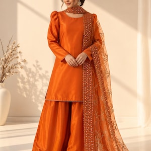 Puede incluir: Un conjunto naranja vibrante de tres piezas que incluye una túnica de manga larga con hombros abullonados, pantalones anchos y una dupatta de organza transparente. La dupatta presenta bordados dorados y espejos.