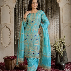 Könnte beinhalten: Ein leuchtend türkisfarbenes Salwar-Kameez-Set mit aufwendigen Goldstickereien und kleinen blauen Akzenten. Die langärmelige Tunika hat einen Rundhalsausschnitt und die passende Hose ist an den Knöcheln gerafft. Ein passender Dupatta mit Goldbesatz liegt über den Schultern.