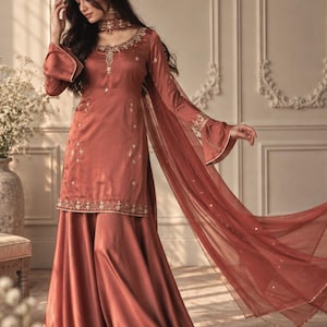 Könnte beinhalten: Ein rostfarbenes Satin-Ensemble bestehend aus einer langärmeligen Tunika mit ausgestellten Ärmeln, einem passenden bodenlangen Rock und einer transparenten Dupatta. Die Tunika ist mit goldener Stickerei am Ausschnitt und Saum verziert. Der Text SAANJH ist sichtbar.