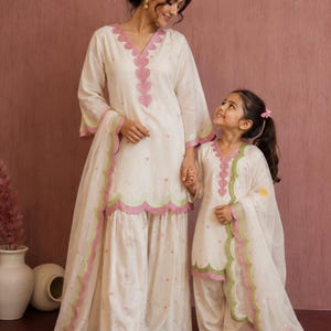 May include: Matching off-white kurta sets featuring scalloped edges in pink and green. The tunics have V-necklines adorned with a vertical row of pink heart-shaped appliqués. Each set includes wide-leg trousers and a sheer dupatta with scalloped trim.