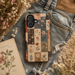 May include: A rectangular phone case featuring a patchwork design of various floral illustrations in muted tones of blue, red, orange, and cream. The case has a black camera cutout area at the top left corner and a smooth, matte-textured surface finish.
