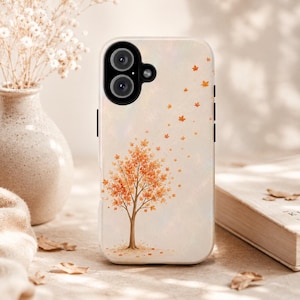 May include: A smartphone case with a cream-colored background featuring a central illustration of a tree with orange and yellow autumn leaves. Some leaves appear to be blowing away from the tree toward the top right corner. The case has a black camera cutout.