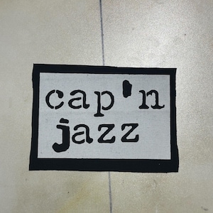Puede incluir: Un parche rectangular de tela con fondo blanco y un borde negro grueso. El texto 'cap'n jazz' está impreso en el centro con una fuente negra en negrita estilo plantilla. El parche tiene una textura tejida plana y bordes rectos y limpios.