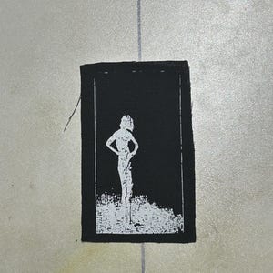 May include: A rectangular black fabric patch featuring a white screen-printed graphic of a person standing with hands on hips. The design has a distressed, high-contrast aesthetic with a thin white border framing the central figure.