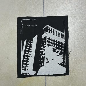 Puede incluir: Un parche de tela rectangular en blanco y negro con un estampado gráfico de alto contraste de un edificio alto. El diseño incluye el texto "i hate myself" en la esquina superior izquierda, escrito en una tipografía sencilla y en minúsculas.