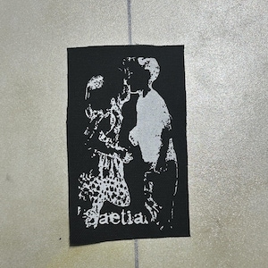 Puede incluir: Un parche rectangular de tela negra con un gráfico serigrafiado en blanco de alto contraste que muestra a dos figuras frente a frente. El diseño incluye la palabra "Gaetia" en una fuente gótica estilizada en la esquina inferior izquierda.