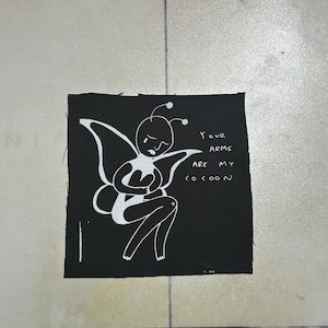 Puede incluir: Un parche de tela negro cuadrado con una ilustración serigrafiada en blanco de una figura alada con antenas, que parece llorar mientras se sujeta el pecho. El texto "YOUR ARMS ARE MY COCOON" está impreso en blanco a la derecha.