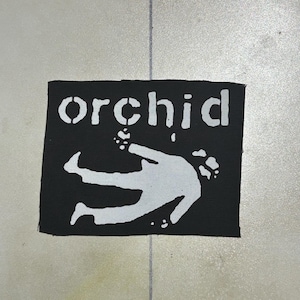 Puede incluir: Un parche rectangular de tela negra con el texto blanco "orchid" en una fuente estilo plantilla sobre una ilustración gráfica blanca de una persona en posición boca abajo con los brazos extendidos, en un diseño plano de alto contraste.