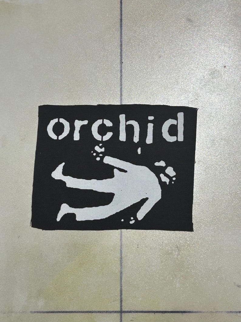 May include: A rectangular black fabric patch featuring the white text "orchid" in a stencil-style font above a white graphic illustration of a person in a prone position with arms extended, rendered in a high-contrast, flat, two-tone design.