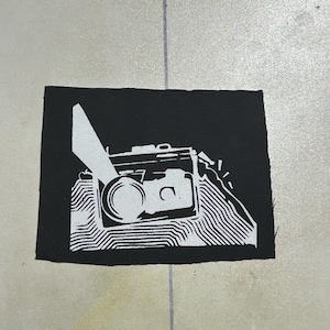 Puede incluir: Un parche rectangular de tela negra con un gráfico serigrafiado en blanco de una cámara. El diseño muestra el cuerpo de una cámara con un objetivo y un flash, rodeado de líneas blancas estilizadas que crean un efecto abstracto y texturizado.