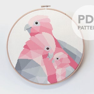 May include: Cross stitch pattern featuring a pink and gray geometric design of two pink cockatoos. The pattern is in a wooden embroidery hoop. The text "PDF PATTERN" is in the upper right corner of the image.