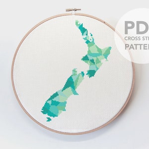 May include: A cross stitch pattern of New Zealand in shades of green and blue, displayed in a wooden embroidery hoop. The pattern is titled "PDF CROSS STITCH PATTERN".
