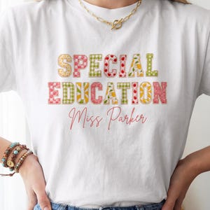 Könnte beinhalten: Ein weißes kurzärmeliges T-Shirt mit dem Schriftzug "SPECIAL EDUCATION" in Blockbuchstaben mit verschiedenen bunten Mustern wie Vichy-Karo, Blumen- und Fruchtprints, gefolgt von dem kursiven Text "Miss Parker" in Korallenrot.