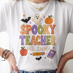 以下が含まれることがあります： SPOOKY TEACHER MS. EMMAという文字がプリントされた白いTシャツ。ゴースト、カボチャ、コウモリ、リンゴ、鉛筆、本が描かれており、オレンジ、紫、緑のチェック柄や様々なパターンが施されたカラフルなグラフィックデザインが特徴です。