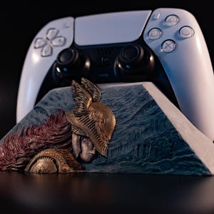 May include: A white wireless game controller rests behind a triangular stand featuring a relief sculpture of a figure with long flowing hair wearing a winged helmet. The sculpture has bronze and blue-grey tones with intricate carved details.