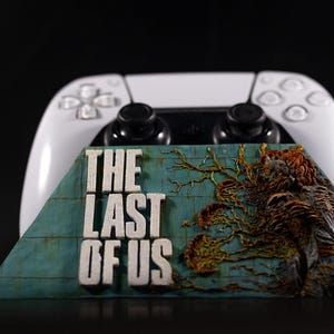 May include: A white video game controller positioned behind a trapezoidal display plaque featuring the text THE LAST OF US in white lettering, set against a teal background with a textured, organic, orange and brown fungal growth pattern.