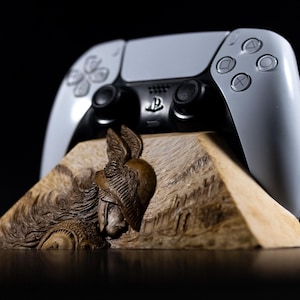 Op de afbeelding: Een wit en zwart draadloze gamecontroller rustend op een lichtgekleurde houten standaard. De voorkant van de standaard bevat een gedetailleerde houtsnijwerk van een krijger met een gevleugelde helm, met complexe texturen en lijnen in het hout.