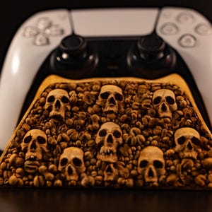 Könnte beinhalten: Ein weißer Videospiel-Controller steht hinter einem dreieckigen Holzständer, der mit einer texturierten Oberfläche aus zahlreichen kleinen, beigen, realistischen Totenkopf-Nachbildungen zwischen vielen braunen Kaffeebohnen verziert ist.
