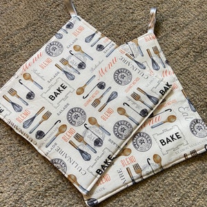 May include: Two beige kitchen pot holders with a white background and a repeating pattern of kitchen utensils, the words "Bake", "Blend", "Culinaire", and "Market Fresh" in black and orange text.