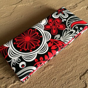May include: A rectangular, folded fabric case with a floral pattern. The design features red, white, and black flowers and leaves. The edges are trimmed with red. The case is likely a glasses case or a similar accessory.