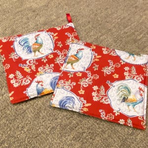 May include: Two red square pot holders featuring a rooster design. Each pot holder has a white oval frame with a rooster illustration. The background is a floral pattern. Each has a red loop for hanging.