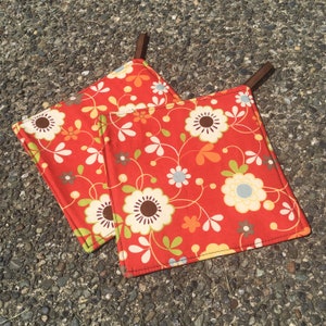 May include: Two red pot holders with a floral pattern in shades of orange, yellow, and white. The pot holders have brown fabric loops for hanging.