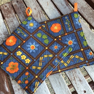 Puede incluir: Dos agarraderas de olla azules y marrones con un diseño floral y de libélula. Las agarraderas tienen un ribete naranja y están hechas de tela.