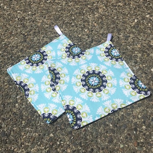 Puede incluir: Dos agarraderas de olla de color azul y blanco con ribete blanco y un diseño verde y azul marino.