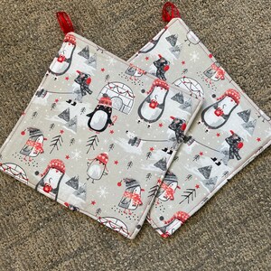 Puede incluir: Dos agarraderas de olla de algodón gris y blanco con un diseño de pingüino y escena de invierno. Las agarraderas tienen lazos de colgar rojos.