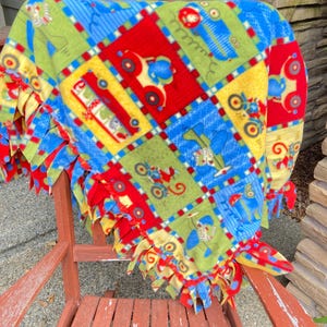 May include: A colorful fleece blanket with a patchwork design featuring cars, motorcycles, and airplanes. The blanket is primarily red, blue, green, and yellow, with a fringed edge. The blanket is draped over a wooden chair.