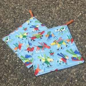 May include: Two square pot holders with a light blue background featuring a pattern of dogs ice skating. The dogs are wearing scarves and sweaters in various colours. Each pot holder has a brown loop.