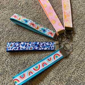 May include: A collection of keychains with colorful patterned wrist straps. Designs include butterflies, rainbows, and suns. Each keychain has a metal clasp for attaching keys.