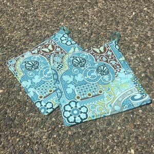 May include: Two teal blue and green patterned pot holders with brown accents. The pot holders have a loop handle on each side.