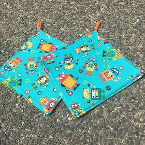 Może przedstawiać: Dwa turkusowe uchwyty do garnków z wzorem robotów. Roboty są żółte, czerwone, niebieskie i zielone. Uchwyty do garnków mają brązowe skórzane pętle do zawieszenia.