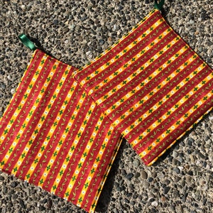 May include: Two square pot holders with a red, yellow, and green floral stripe pattern. Each pot holder has a green ribbon loop for hanging. The pot holders are made of fabric and are designed to protect surfaces from heat.