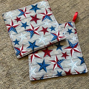 May include: Two red, white, and blue star-patterned pot holders with red hanging loops. The pot holders are on a beige carpet.