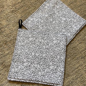 May include: Two grey and white leaf-patterned pot holders with black hanging loops. The pot holders are square and made of fabric. The leaf pattern is a repeating design of white leaves with black outlines.