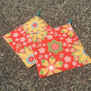 Puede incluir: Dos agarraderas de olla de tela con estampado floral rojo y amarillo con lazos de suspensión de color turquesa.