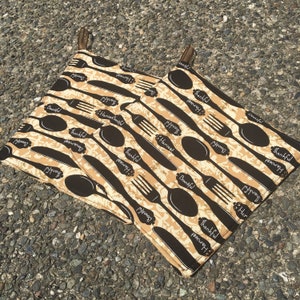 May include: Two brown and beige pot holders with a repeating pattern of black forks, spoons, and knives. The pot holders have the words "Thankful", "Harvest", and "Bountiful" printed on them.
