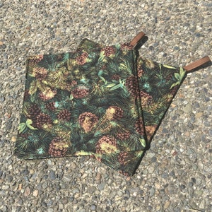 May include: Two brown and green pot holders with a pine cone and evergreen pattern. The pot holders have brown leather straps.
