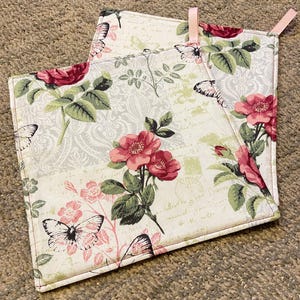 May include: Two square pot holders with a floral and butterfly pattern. The design features pink roses, green leaves, and butterflies on a cream background with script writing. Each pot holder has a pink loop for hanging.