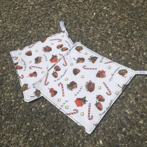 May include: Two white pot holders with a Christmas theme. The pot holders feature a red and white candy cane, a red and green holly sprig, a gold star, and a chocolate-covered strawberry. The text on the pot holders reads "Tis the season... to eat chocolate." and "la la la la... la la la la!"